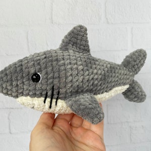 Puede incluir: Un juguete de peluche de tibur&oacute;n gris tejido a crochet con detalles blancos alrededor de la boca y un ojo negro.