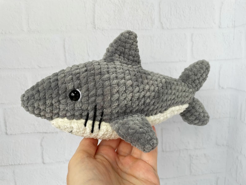 Puede incluir: Un juguete de peluche de tibur&oacute;n gris tejido a crochet con detalles blancos alrededor de la boca y un ojo negro.