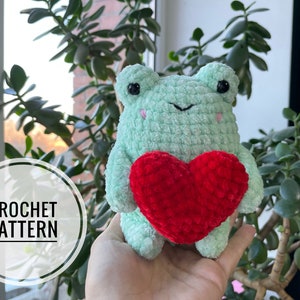 May include: A crocheted green frog holding a red heart. The frog has black eyes and a small smile. The heart is made of red yarn and has a textured surface.
