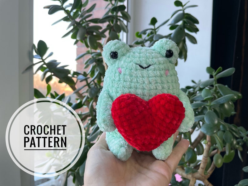A crocheted green frog holding a red heart. The frog has black eyes and a small smile. The heart is made of red yarn and has a textured surface.