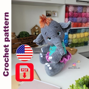 Może przedstawiać: Ręcznie robiona szara szydełkowa lalka słonia z pomarańczowymi włosami, w turkusowej górze i różowej spódnicy. Obraz zawiera ikonę PDF, amerykańską flagę i tekst "Crochet pattern". Lalka siedzi na białej powierzchni.