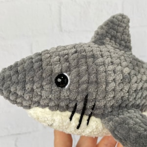 Puede incluir: Juguete de peluche de tibur&oacute;n gris tejido a crochet con ojos negros y vientre blanco. El tibur&oacute;n tiene una boca cosida con l&iacute;neas negras.