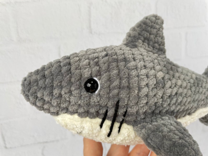 Puede incluir: Juguete de peluche de tibur&oacute;n gris tejido a crochet con ojos negros y vientre blanco. El tibur&oacute;n tiene una boca cosida con l&iacute;neas negras.