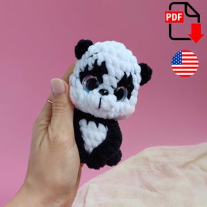 May include: A handmade crochet panda bear with black and white yarn. The panda has large, blue eyes and small black ears. The panda is held in a hand against a pink background. A PDF icon and an American flag are in the background.