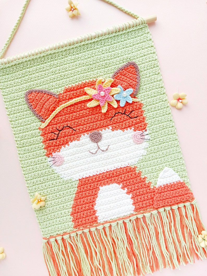 Op de afbeelding: Een gehaakte wandhanger met een vos met een bloemenkrans. De vos is oranje met een witte buik en een lichtgroene achtergrond. De wandhanger heeft een franje van lichtgroen en oranje garen.