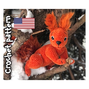May include: A crocheted orange squirrel plush toy with a fluffy tail, sitting on a branch with snow in the background. The toy has black eyes and a black nose.