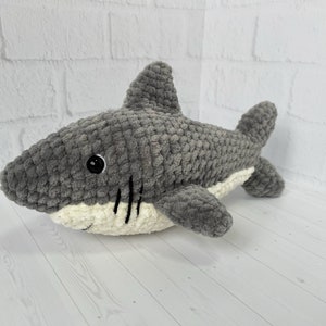 Puede incluir: Un juguete de peluche de tibur&oacute;n de crochet gris y blanco. El tibur&oacute;n tiene la barriga blanca y los ojos negros.