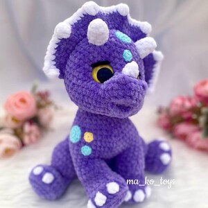 May include: Purple crocheted triceratops stuffed animal with white accents and blue and yellow spots. The toy is sitting on a white surface.