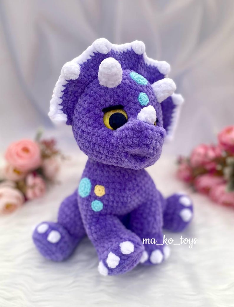 May include: Purple crocheted triceratops stuffed animal with white accents and blue and yellow spots. The toy is sitting on a white surface.