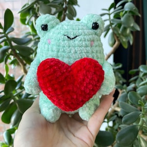 May include: A green crocheted frog holding a red crocheted heart. The frog has black eyes and a pink nose.