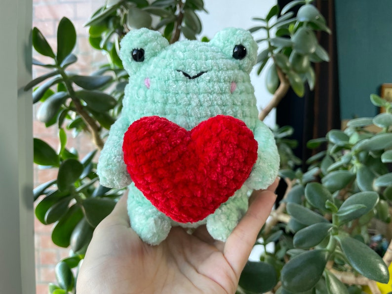 May include: A green crocheted frog holding a red crocheted heart. The frog has black eyes and a pink nose.