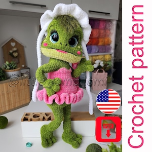 Könnte beinhalten: Eine grüne gehäkelte Froschpuppe mit großen blauen Augen, rosa Wangen und einer weißen Haube, die ein rosa Rüschenkleid trägt. Das Bild enthält den Text Crochet pattern, ein US-Flaggen-Symbol und ein rotes PDF-Download-Symbol rechts.