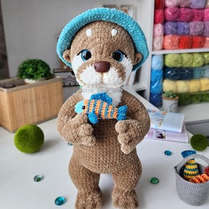 Front view of crocheted otter holding a blue and orange striped fish, wearing a turquoise hat. Detailed and soft amigurumi made with plush yarn for a cute DIY gift.