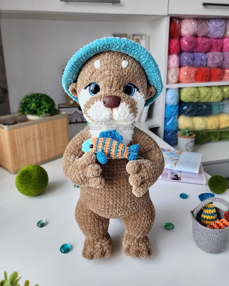 Front view of crocheted otter holding a blue and orange striped fish, wearing a turquoise hat. Detailed and soft amigurumi made with plush yarn for a cute DIY gift.