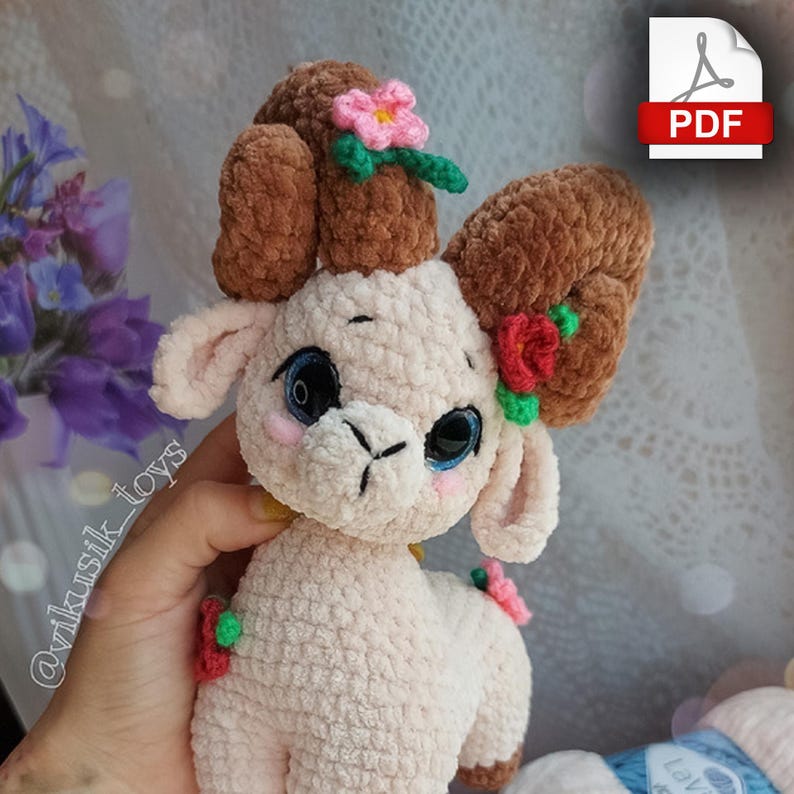 May include: A crocheted plush toy lamb with brown curly horns, pink and red flowers, and green leaves. The lamb has a white body and blue eyes.