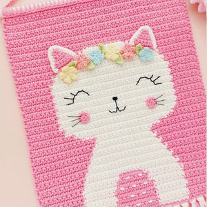 Peut inclure: Une tapisserie murale en crochet rose avec un chat en crochet blanc portant une couronne de fleurs. Le chat a un nez rose et des moustaches blanches. La tapisserie murale est orn&eacute;e de franges blanches.