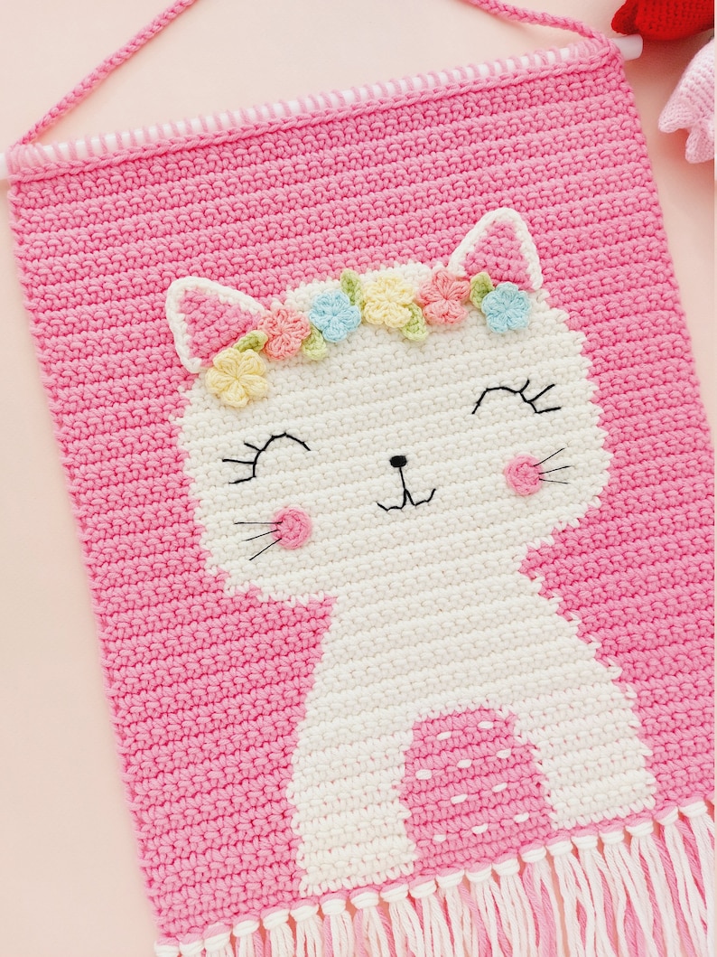Peut inclure: Une tapisserie murale en crochet rose avec un chat en crochet blanc portant une couronne de fleurs. Le chat a un nez rose et des moustaches blanches. La tapisserie murale est orn&eacute;e de franges blanches.