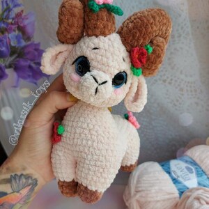 May include: A crocheted plush toy lamb with brown curly horns, pink fur, and green and red flower accents. The lamb is holding a yellow flower in its mouth. The toy is made with soft yarn and has a sweet, whimsical expression.