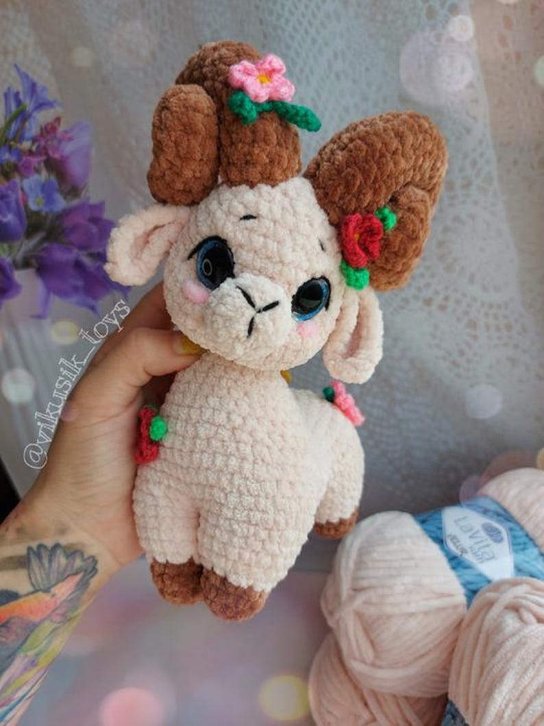 May include: A crocheted plush toy lamb with brown curly horns, pink fur, and green and red flower accents. The lamb is holding a yellow flower in its mouth. The toy is made with soft yarn and has a sweet, whimsical expression.
