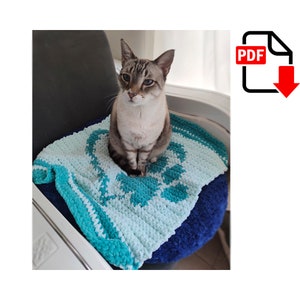 May include: A teal and light blue crocheted cat bed with a cat sitting on it. The cat bed has a flower design in the centre.