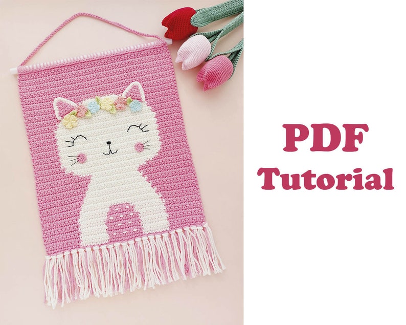 Peut inclure: Une suspension murale en crochet rose avec un motif de chat blanc et un bandeau floral. Le chat a un nez rose et des moustaches blanches. La suspension a une frange blanche en bas. Le texte "Tutoriel PDF" est en lettres roses sur fond blanc.