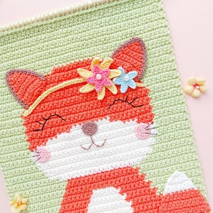 Op de afbeelding: Een gehaakte wandhanger met een vos met een bloemenkroon. De vos is oranje met witte accenten en glimlacht. De achtergrond is groen en de hanger heeft een franje rand.