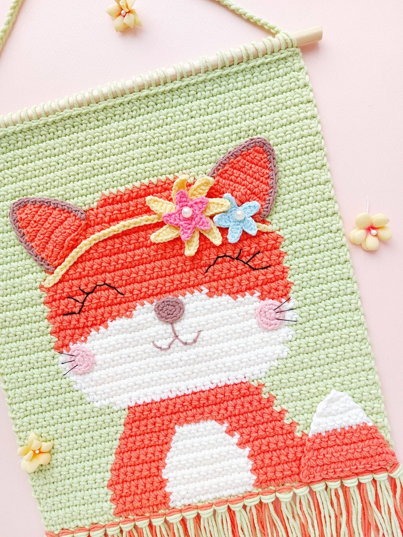 Op de afbeelding: Een gehaakte wandhanger met een vos met een bloemenkroon. De vos is oranje met witte accenten en glimlacht. De achtergrond is groen en de hanger heeft een franje rand.