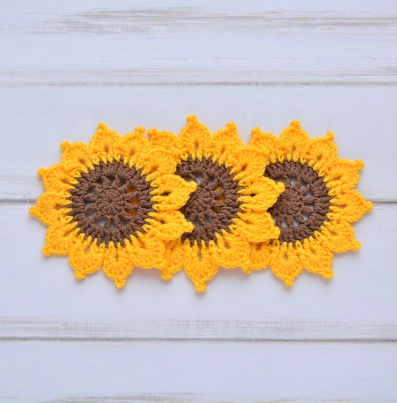 May include: Three crocheted sunflower coasters in yellow and brown. The coasters are arranged in a row, with the centers of the sunflowers facing the viewer.
