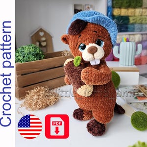 Könnte beinhalten: Ein handgehäkelter brauner Biber mit blauer Mütze, der einen Baumstamm mit einem grünen Blatt hält. Der Biber hat weiße Zähne und blaue Augen. Das Bild enthält den Text "Crochet pattern" und ein PDF-Download-Symbol.