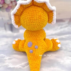 May include: A crocheted yellow triceratops dinosaur toy with a white frill and purple spots. The toy is sitting on a white surface.
