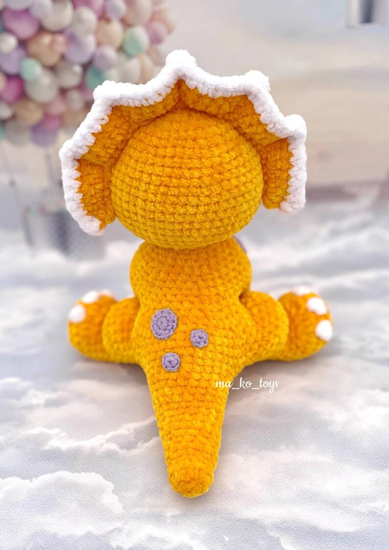 May include: A crocheted yellow triceratops dinosaur toy with a white frill and purple spots. The toy is sitting on a white surface.