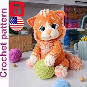 Peut inclure: Une poupée chat en crochet orange et blanche avec des yeux bleus et un nez rose, tenant une pelote de laine verte. La poupée a des marques rayées et est assise. L'image comprend le texte "Crochet pattern".