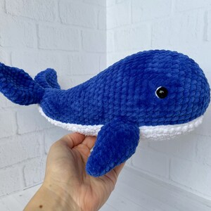 May include: A blue crocheted whale plush toy with a white belly and a black eye. The whale is lying on its side with its tail curled up.