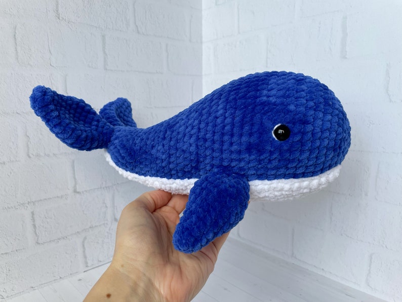 May include: A blue crocheted whale plush toy with a white belly and a black eye. The whale is lying on its side with its tail curled up.