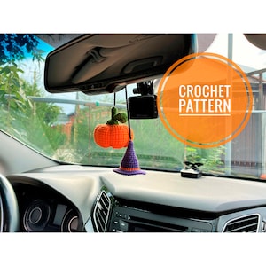 May include: Crocheted Halloween car charm featuring an orange pumpkin and a purple witch hat with orange trim. The charm hangs from a black string, with the text "CROCHET PATTERN" in an orange circle.