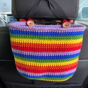 May include: A rainbow striped crocheted car seat organizer with two wooden buttons. The organizer is hanging from the headrest of a car seat.