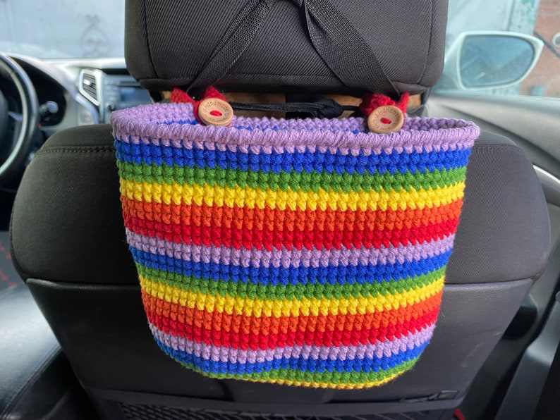 May include: A rainbow striped crocheted car seat organizer with two wooden buttons. The organizer is hanging from the headrest of a car seat.