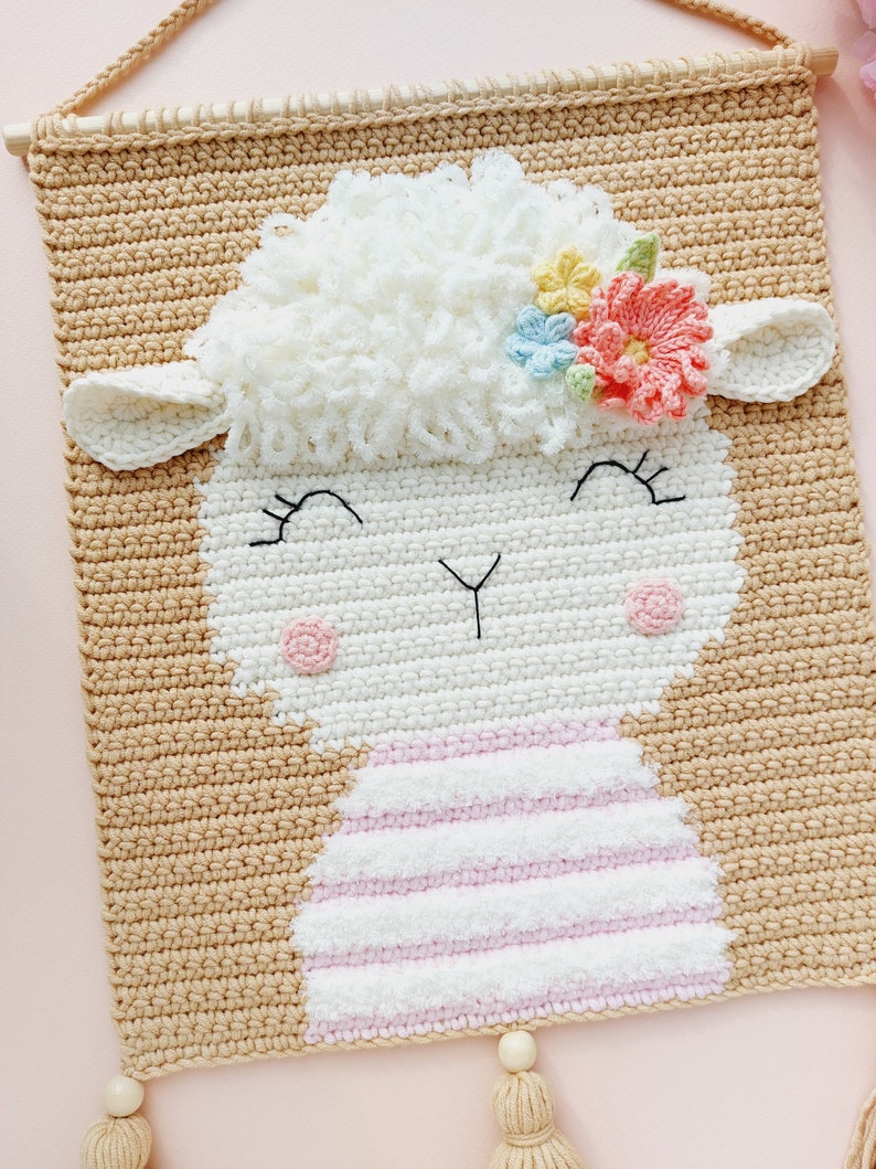 May include: A crocheted wall hanging featuring a white sheep with a pink and white striped dress and a flower crown with pink, blue, and yellow flowers. The sheep has a smiling face with black eyes and a black nose.