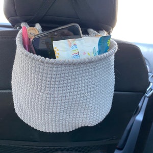 May include: A gray crocheted hanging basket with a phone, a pen, and a package of snacks inside. The basket is hanging from the headrest of a car seat.