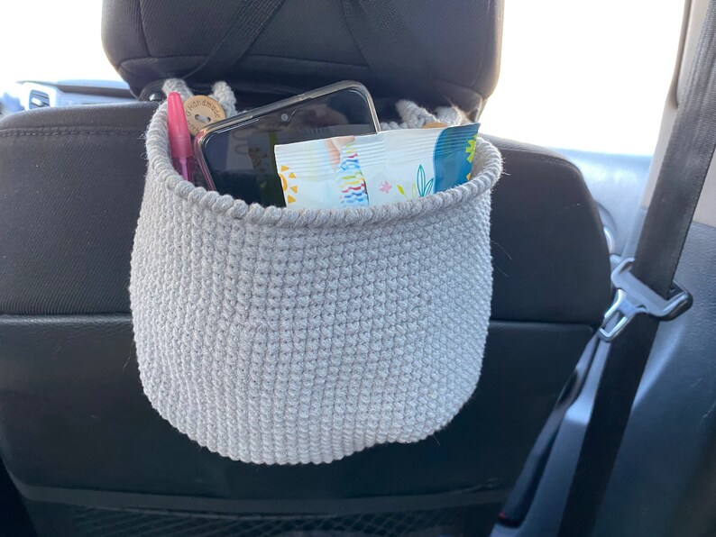 May include: A gray crocheted hanging basket with a phone, a pen, and a package of snacks inside. The basket is hanging from the headrest of a car seat.