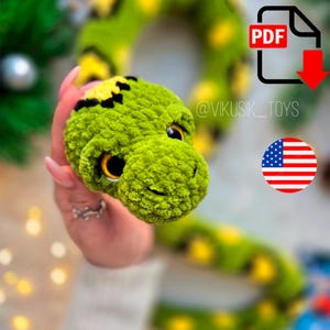 Puede incluir: Un juguete de peluche de serpiente verde hecho a mano con detalles amarillos y negros. La cabeza de la serpiente está enfocada, mostrando ojos detallados y una boca sonriente. El juguete se sostiene en una mano, con un fondo borroso.