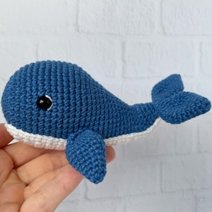 May include: A crocheted blue whale toy with white accents and black eyes. The whale is lying on its side with its tail curled up.