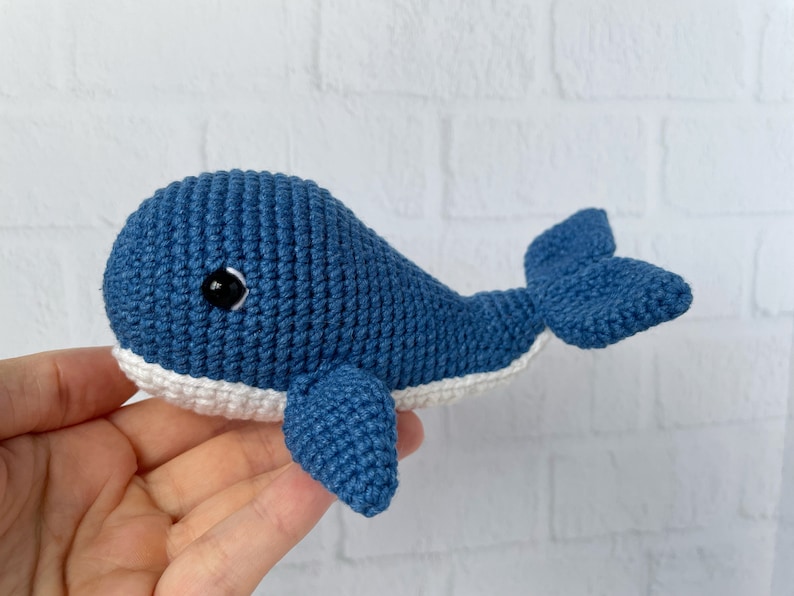 May include: A crocheted blue whale toy with white accents and black eyes. The whale is lying on its side with its tail curled up.