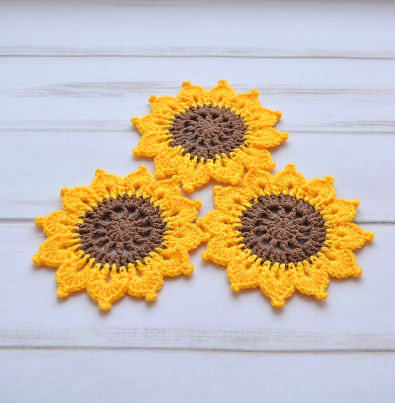 May include: Three crocheted sunflower coasters in yellow and brown. The coasters are arranged in a triangular shape on a white wooden surface.