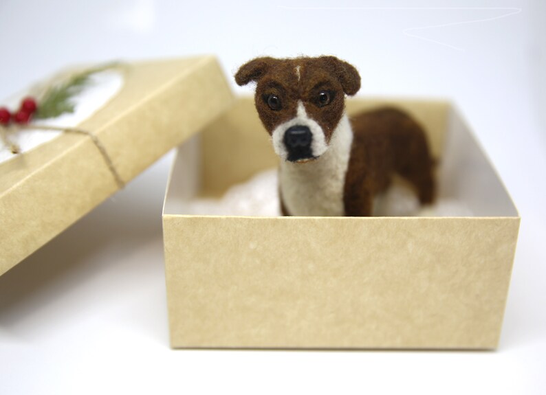 May include: A brown and white dog figurine inside a tan gift box. The dog has a white chest and face with brown markings. The box is open, revealing the dog and a white interior. A sprig of greenery and red berries adorn the box.