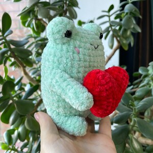 May include: A green crocheted frog holding a red crocheted heart. The frog has black eyes and a pink nose.