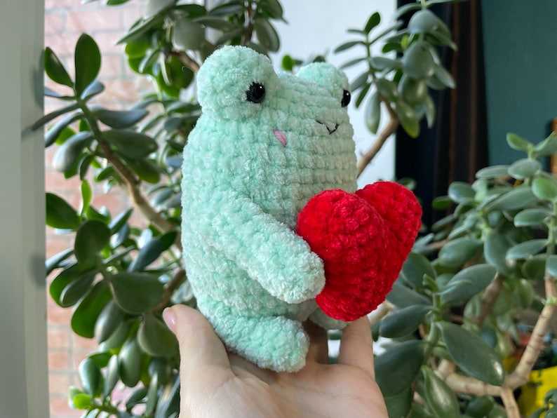 May include: A green crocheted frog holding a red crocheted heart. The frog has black eyes and a pink nose.
