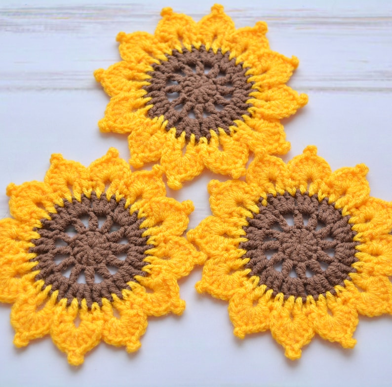 May include: Three crocheted sunflower coasters in yellow and brown. The coasters are made with a detailed pattern and have a raised center.