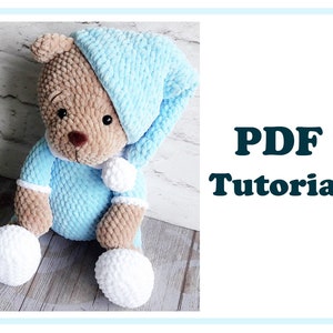 Puede incluir: Un oso de peluche tejido a crochet con un gorro de dormir azul y pijama. El oso está sentado sobre una superficie de madera. El texto "PDF Tutorial" está en el fondo.