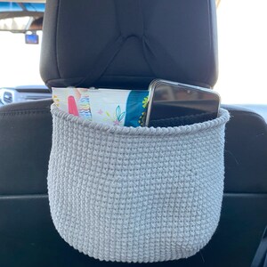 May include: A gray crocheted car seat organizer hanging from the headrest of a car seat. The organizer is filled with a package of wipes and a smartphone.